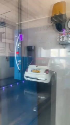 A white Fiat 500 is being washed in a car wash. The car is positioned on a track and is moving through the wash. Water jets are spraying the car from the sides. There are blue and purple lights in the car wash. The car has a Dutch license plate that reads "51-LZP-6". The car wash is branded with "Aral SuperWash". The video is captured from behind a glass partition, showing reflections of the environment.