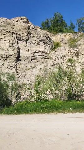 A handheld video pans across a sun-drenched, rugged cliff face. The cliff is composed of layered rock in shades of tan, beige, and a greenish-blue hue, with visible cracks and fissures. Sparse green vegetation, including bushes, trees, and yellow wildflowers, grows at the base of the cliff and on its upper slopes. A dirt path runs along the bottom of the frame, partially obscured by the vegetation. The sky above is a clear, bright blue. The video is shot from a low angle, looking upwards towards the imposing cliff.