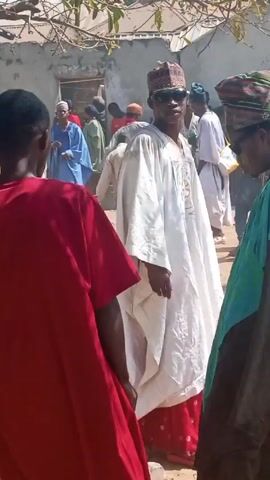 The video shows a crowd of people gathered outdoors, likely in a village or small town setting. Many are wearing traditional West African clothing, including long flowing robes called boubous, and various types of hats. The ground appears to be dirt or sand. Some people are holding water bottles. The overall atmosphere seems festive, possibly a celebration or gathering. A low-resolution camera and shaky filming prevent detailed identification of individual features, though most of the people seem to be male.