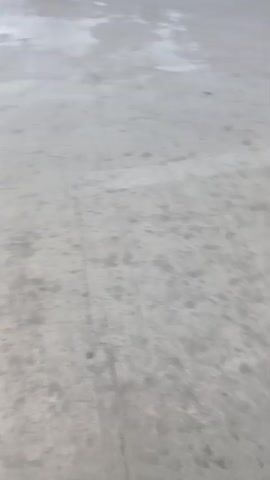 The video shows a ground-level view of a concrete surface. A dark line runs down the middle of the frame. In one section, a faint yellow line is visible parallel to the dark line. The concrete appears wet or damp in places, creating lighter patches. The camera angle is slightly tilted, giving a sense of movement as if someone is walking or riding. There are no people or objects clearly visible in the frame. The environment appears to be an outdoor paved area, possibly a road, parking lot, or pathway. The lighting is overcast.