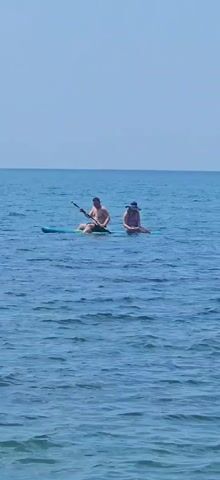 A man and a woman are paddleboarding together on a calm blue ocean under a clear sky. The man is in front, facing forward and holding a paddle. The woman is behind him, facing forward and wearing a hat. They are on a single paddleboard, with the man sitting and the woman sitting behind him. The water is calm with gentle ripples.