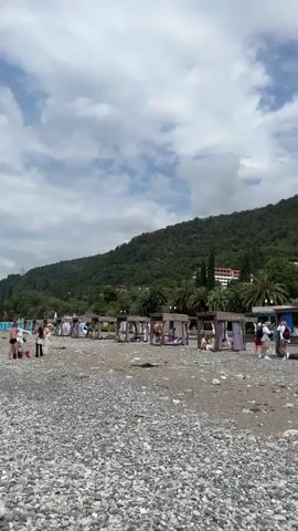 The video pans across a pebble and sand beach with a lush green mountain in the background. Several wooden beach cabanas with purple curtains are set up along the shore. Some people are visible sitting in or near the cabanas. A small wooden building, possibly a shop, is situated on the beach. Further down the beach, a fence with surfboards and paddleboards is visible. Palm trees are scattered throughout the scene. The sky is partly cloudy.