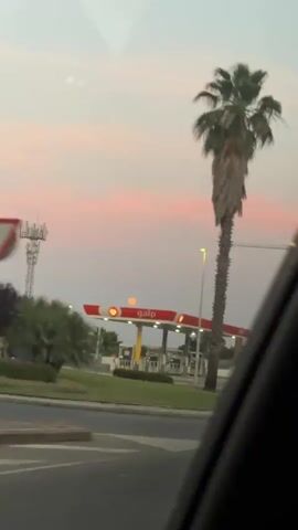 The video appears to be taken from inside a moving vehicle, possibly a car, as it drives on a road in what looks like a commercial area. The focus is on the right side of the road. There is a GALP gas station with its red and white canopy and gas pumps visible. A tall palm tree stands near the roundabout in front of the gas station. Further down, an advertisement billboard for AMK RENT, which rents vans, is visible, including a white van image, phone numbers, and a website address. The sky is a pale pink color with some clouds, suggesting it might be near sunrise or sunset.  There's also a construction crane visible in the distance. No people are clearly visible.