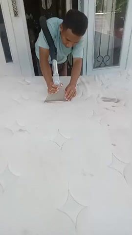A young man in a light blue t-shirt and dark shorts is cleaning a white mattress with a wet vacuum cleaner. He is holding the vacuum attachment with both hands and moving it across the surface of the mattress. The mattress appears to be dirty with visible stains. The scene is outdoors, in front of a white door or window frame, with the mattress laid out on a white surface. Sunlight is visible, suggesting it's daytime.
