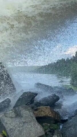 A wave crashes against rocks on a shore. The water is a murky brown and white as it churns and sprays. The rocks are dark gray and appear wet and covered in moss in some areas. The sky is a clear blue with a few wispy clouds. The overall scene is one of natural power and the raw force of the ocean.