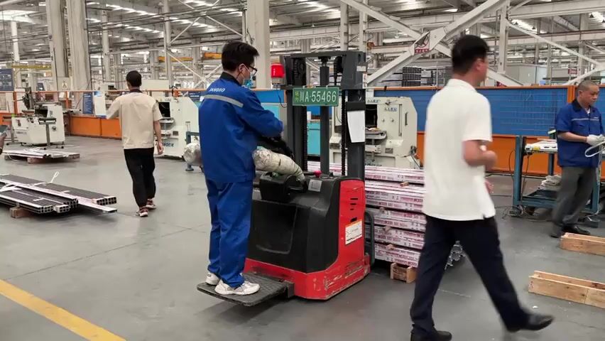 A factory worker wearing a blue jumpsuit and a face mask is operating a red and black electric forklift inside a large factory. The forklift is carrying a stack of long, silver, wrapped metal pieces. Other workers are present, some walking around and others standing nearby. One person is holding a smartphone and another is wearing a white polo shirt. The factory floor is concrete and well-lit with industrial machinery in the background.  The forklift has a license plate that reads "苏A-55466".  A red light is blinking on top of the forklift.