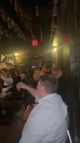 The video shows a dimly lit bar with a lively atmosphere, where patrons are singing along to live piano music. The interior has a rustic feel with exposed brick walls and decorative objects hanging from the ceiling. A pianist plays on a small stage, visible on a screen, while the crowd raises their arms, drinks, and cell phones in unison, creating a cheerful and energetic scene.