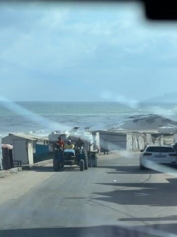 The video shows a view from inside a vehicle looking out through a windshield. A blue tractor pulling a trailer with large white water tanks is driving down a paved road. Three people are visible on the tractor and trailer; one person is driving the tractor, another is sitting on the trailer behind the tractor, and a third person is sitting on the front of the trailer next to the driver. The tractor and trailer are moving towards the right of the frame. A white car is parked or driving on the right side of the road. In the background, there are several makeshift structures, possibly tents or shacks, lining the road. Beyond the structures is a sandy beach and the ocean under a cloudy sky. A young child wearing a red shirt and blue shorts is running across the road towards the right of the frame, away from the tractor and trailer, and in the general direction of the white car. The child appears to be barefoot. The lighting suggests it is daytime.