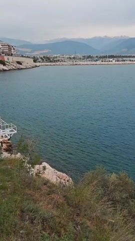 The video presents a serene, panoramic view of a tranquil bay or sea, slowly panning from left to right. The camera is positioned on a slight elevation overlooking the water.

In the foreground, the immediate coastline features rugged, light-colored rocks interspersed with sparse green and brown bushes and vegetation. The water directly below the camera appears relatively clear, revealing submerged rocks near the shore before deepening to a dark blue-green hue further out.

The midground is dominated by the calm expanse of water, showing gentle ripples across its surface.

In the distant background, a range of hazy mountains stretches across the horizon, some with faint snow patches visible. As the video begins, on the far left across the bay, a coastal town or settlement is visible, featuring numerous buildings and a prominent marina with what appear to be docked boats. As the camera pans right, this urban area recedes from view, giving way to the open water and continuous mountain range.

The sky is overcast or partly cloudy, casting a soft, diffused light over the landscape, suggesting either an early morning, late afternoon, or simply a cloudy day. No people or visible text are present in the video frame. The overall atmosphere is one of peacefulness and natural beauty in a coastal setting.