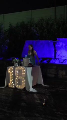 A person is standing behind a table covered with a white cloth, where a cake and some decorative plants are placed. Large illuminated numbers "18" are in front of the table. The scene takes place outdoors at night, with plants and a wall in the background. Blue lights illuminate a rock formation behind the table. A person in a light blue shirt and white pants is standing to the right of the frame, facing the table. The ground appears to be wet. The illuminated numbers and the general atmosphere suggest a celebration, possibly a birthday. The audio indicates that it is close to midnight.