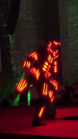 The video shows a nighttime outdoor performance on a stage illuminated by red and green lights. Two performers are visible. The performer on the left is wearing a black costume with white stripes and appears to be dancing. The performer on the right is wearing a costume made of black material and illuminated with bright red LED lights in a geometric pattern. Another performer in the background is wearing a green LED illuminated costume and is also dancing. In the far background, a stone wall can be seen, suggesting a historical or outdoor venue. Several audience members are dimly visible in the background, observing the performance. The overall scene depicts a dynamic and visually striking performance, likely involving synchronized dancing and light effects.