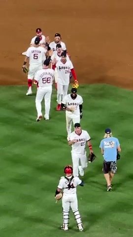 A group of baseball players wearing white uniforms with red trim walk across a green field. One player in the foreground, wearing a catcher's mask and gear, high-fives another player. Several players are wearing the word "Angels" across their chests. Behind them, a person in a blue shirt and patterned shorts walks in the same direction. The field appears to be part of a baseball stadium, with dirt visible in the background.