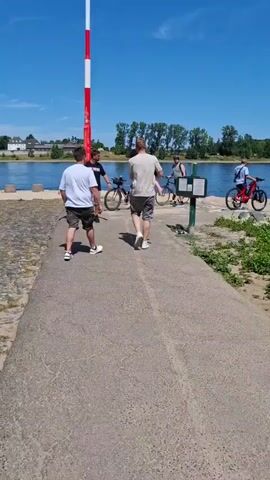 This video captures a sunny day scene by a river. In the foreground, a paved path leads towards a body of water, with people walking and biking. A red and white striped pole stands tall on the left, possibly a navigational marker. On the right, lush green bushes line the sandy bank. A white passenger boat is docked near the shore, with a few people boarding or disembarking. Further in the distance, a larger vessel, possibly a cargo ship, is visible on the river. The sky is a clear blue with a few wispy clouds, and the overall atmosphere is one of leisure and outdoor activity.