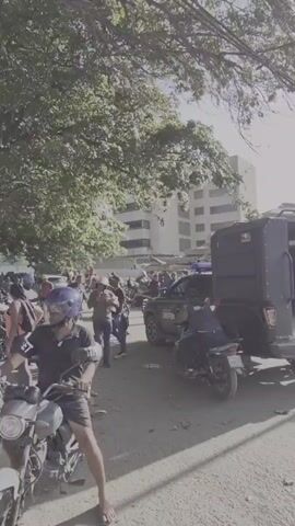 The video shows a street scene in front of a collapsed building. Several people are present, some on motorcycles and others walking. A black vehicle with a cage-like structure on the back, possibly a police vehicle, is parked on the street. Another black SUV is also visible. There is debris from the collapsed building in the background, and a bulldozer is present near the wreckage. The overall scene appears to be one of a disaster or accident response.