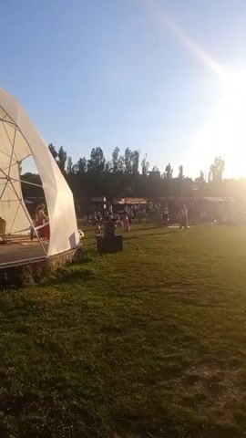 The video shows an outdoor gathering of people in a grassy area during sunset. The sun is very bright and positioned in the upper right corner of the frame, casting lens flares and creating a hazy effect. In the foreground, there is a patch of mown grass. To the left, a white tent-like structure is partially visible. In the middle ground, a crowd of people are scattered across the grass, many sitting or lying down. Some appear to be near food stalls or tents. In the background, a line of trees and some structures are silhouetted against the bright sky. The overall atmosphere appears relaxed and communal, with people enjoying the evening.