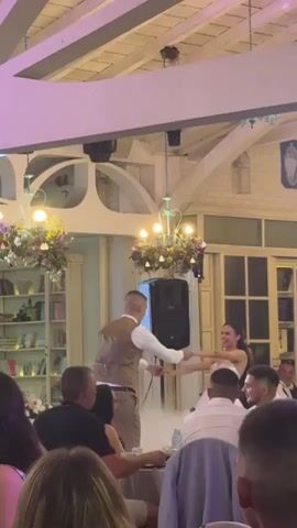 In a wedding reception hall, a bride and groom dance while a sparkler effect erupts around them. The bride, wearing a white wedding dress, smiles joyfully as she twirls with her groom, who is dressed in a tan vest and white shirt. Their dance takes place on a raised platform, shrouded in a light fog, with a large speaker positioned behind them.

The hall's interior is visible, featuring exposed wooden beams on the ceiling with colorful lights projected onto them. Two ornate chandeliers, adorned with floral arrangements, hang from the ceiling. In the background, bookshelves filled with books line a wall, and large windows with white trim offer a glimpse of the outside.

A diverse group of guests are seated at tables, watching the couple dance. Some guests are looking towards the couple, while others are engaged in conversation. The atmosphere appears celebratory and joyful. The image captures a dynamic moment of a wedding celebration, highlighted by the special effects and the couple's affectionate interaction. The context of Krepost, Bulgaria, suggests a European wedding setting.