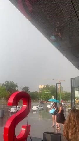 The video captures an outdoor scene on a wet, overcast day, possibly during or immediately after a rain shower. Two women are visible in the foreground, one holding a blue umbrella over both of them. They are standing on a wet paved area, with reflections of the sky and buildings visible on the ground. Behind them, a large red sculptural element, possibly a stylized number or letter, is prominently displayed. In the background, trees, streetlights, cars, and buildings are visible, suggesting an urban environment. The sky is mostly cloudy, with some patches of lighter sky indicating potential breaks in the clouds. The overall mood appears subdued due to the weather.