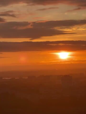 The video captures a vertical panoramic shot of a cityscape at sunset. The sky is dominated by a vibrant orange glow, with dark clouds scattered across the upper portion of the frame. The sun, a bright white orb, is visible just above the horizon, casting a warm light over the entire scene. In the foreground, the silhouetted rooftops of several buildings are visible, appearing as dark, geometric shapes against the bright sky. The rest of the cityscape is shrouded in a hazy, orange mist, making it difficult to discern individual buildings or features. The overall impression is one of a peaceful, serene sunset over a sprawling urban landscape.