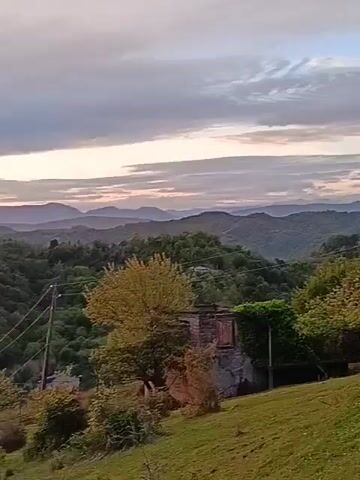 The video pans across a rural landscape at sunset. Lush green hills covered in trees roll into the distance, where deeper, purplish mountains stand under a sky brushed with shades of pink, orange, and gray. The sun is setting, a bright, fiery orb just above the horizon. A few houses are nestled among the hills, some with smoke rising from their chimneys. Closer to the camera, there's a patch of grass and an old, somewhat dilapidated building. Utility poles with wires run across the foreground, cutting through the scene. The overall impression is one of tranquility and scenic beauty as dusk settles over the countryside.