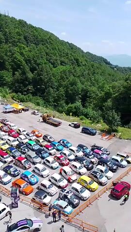 The video pans over a large outdoor gathering of people and many cars. The cars are parked in neat rows in a paved area that appears to be a parking lot. Many of the cars are brightly colored and appear to be sports cars or modified vehicles. Some cars are white, red, blue, yellow, green, and orange. There are also several people walking around the cars, some of whom are gathered in small groups. In the background, there is a large, wooded hill covered in green trees. Several tents and structures are visible, including a large white tent with an arched entrance, and several smaller tents with company logos, such as Red Bull. A tow truck is also visible in the middle ground. The weather appears to be sunny and clear.