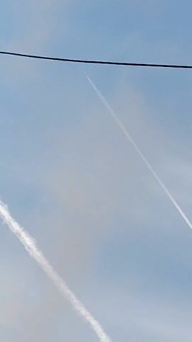 The video shows a sky with contrails from airplanes. There are at least two contrails visible, with one appearing as a clear white line extending across the frame and another shorter, somewhat dispersed contrail below it. In the upper part of the frame, there is a black wire extending horizontally, partially obstructing the view of one of the contrails. The sky is light blue with some wispy clouds. An airplane is visible in the distance, leaving a contrail.