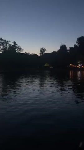 The video captures a serene twilight scene at Lake Chiemsee, near Gstadt am Chiemsee, Germany. The camera pans slowly across the water, reflecting the deepening blue of the sky. Silhouetted against the horizon are trees and a steeple, suggesting a quaint village or town lining the shore. On the right side of the frame, a cluster of lights twinkles, indicating a gathering of buildings, possibly a restaurant or a small harbor, with their reflections shimmering on the water's surface.

The time of day is clearly evening, as the sky transitions from day to night, with the last vestiges of daylight fading. The water is calm, with gentle ripples disturbing its surface, creating a peaceful and tranquil atmosphere. There are no discernible people or specific objects in sharp focus, the scene emphasizes the overall ambiance and the natural beauty of the location. The mood is one of quiet contemplation and the gentle fading of the day. No text is visible in the media.