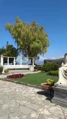 The video shows a sunny outdoor scene. First, it pans across a garden featuring a statue of a seated man in a toga, a green lawn, a white stone colonnade, potted plants, and trees under a clear blue sky. The camera then transitions to a selfie perspective of a young woman with sunglasses on her head. She is wearing a white top. The woman smiles at the camera and walks along a stone-paved path. In the background, people can be seen walking around.