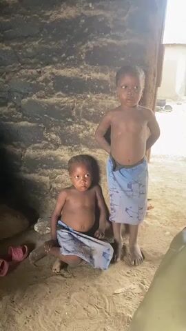 Two young, dark-skinned children stand in the doorway of a hut. The older child is standing, with hands behind her back. The younger child is squatting, leaning against the wall. Both children are wearing cloth wraps around their waists. The wall behind them is dark, and appears to be made of mud or clay. There is sand on the ground. Pink sandals are visible on the left. The light is bright.