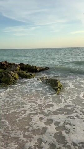 The video captures a serene beach scene with gentle waves lapping at the shore. In the foreground, the sand is wet from the receding tide, with small pebbles visible. Several large, moss-covered rocks jut out from the water on the left side of the frame, with waves breaking around them and creating white foam. The ocean stretches out to the horizon, its surface a mix of turquoise and green hues. The sky above is a pale blue, with scattered wispy clouds. There are no people or animals visible in the video. The overall impression is one of tranquility and natural beauty.