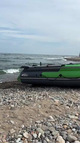 A green and grey inflatable boat rests on a pebbled beach next to a larger blue boat. Waves lap at the shore and the sky is overcast.