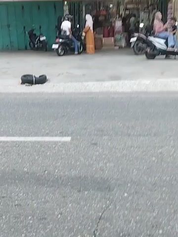 The video shows a street scene in front of a store. There are several motorcycles parked on the sidewalk, and a few people are present. One woman in an orange dress and a headscarf is standing near a motorcycle. Another woman in a pink hijab is sitting on a motorcycle. A man in a white shirt and jeans is on another motorcycle. There is a tire lying on the sidewalk. The store appears to be selling various goods, including what look like bottles of liquid and electronic devices. A sign above the store entrance reads "KREDIT" and "ADMIN". The road in the foreground is asphalt with a white line marking a lane. A red motorcycle is speeding past in the foreground, blurring the image. The overall impression is a bustling commercial area.