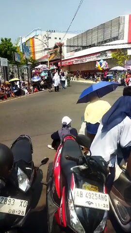 The video shows a street scene with several people, motorcycles, and buildings. In the foreground, two motorcycles are parked. One is black with license plate 2893 XO, and the other is red and white with license plate AB 4925 AHG. A person wearing a white hijab and a dark blue jacket is partially visible sitting on the ground between the motorcycles. Further down the street, more people are gathered, some holding umbrellas, suggesting a sunny day. A few individuals are walking, and there are several parked motorcycles and scooters. Buildings line the street, with some appearing to be commercial establishments. The overall environment seems to be an urban or semi-urban setting.