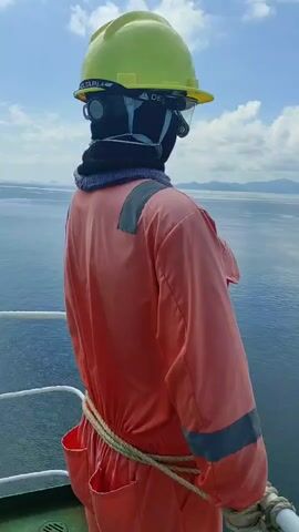 The video features an individual, seen from a slightly elevated side-rear perspective, standing on the deck of what appears to be a ship or boat. The person is dressed in full protective work gear, including a bright yellow hard hat with "DELTAPLUS" visible on it, clear safety glasses, a black balaclava or face mask, and a dark neck warmer. They are wearing a peach-orange long-sleeved jumpsuit with grey reflective strips on the shoulders, and a rope is tied around their waist. White work gloves are also visible.

The individual is looking out over a wide expanse of calm, blue sea. In the distance, several islands or landmasses are visible on the horizon under a partly cloudy sky. The bright lighting suggests it is daytime. A white railing is present in the foreground, indicating the edge of the vessel's deck. The scene portrays a maritime environment, specifically located in Kunak, Malaysia, with the individual seemingly observing the open water and distant land.