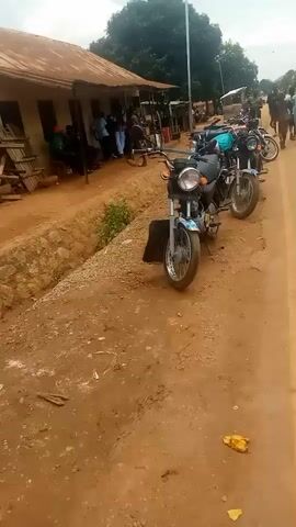 A stationary video captures a dirt road alongside a building with a corrugated metal roof. Several motorcycles are parked in a line on the side of the road, facing away from the camera. Some people are visible in the background, standing near the building or on the road. The scene appears to be an outdoor marketplace or gathering place.
