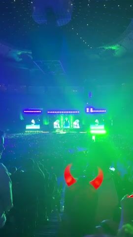 The video shows a concert with a large audience. The stage is illuminated with bright green and purple lights, and screens display images of performers. In the foreground, a person is visible wearing red devil horns. The overall atmosphere appears to be energetic and festive.