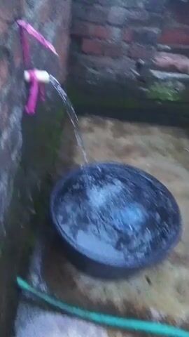 The video shows a close-up shot of a water tap attached to a brick wall. Pink fabric is tied around the tap. Water is flowing from the tap into a large black basin, creating ripples on the surface of the water. The wall is made of red bricks and appears slightly damp. The ground is concrete and also looks wet.