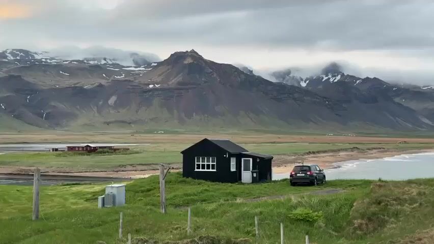 The video captures a wide, static shot of a rugged coastal landscape in what appears to be Búðir, Iceland. In the midground, a small, black single-story house with white window frames and a white front door stands on a grassy knoll. A dark-colored SUV is parked on the grass next to the house, facing the ocean. To the right of the house, a calm sea stretches out, meeting a sandy and rocky shoreline. In the foreground, green grass slopes gently, featuring a few wooden posts and a small, light-colored utility box.

The background is dominated by a majestic range of dark, jagged mountains. Many of the peaks are partially covered in snow, and low-hanging, grey clouds obscure their tops and higher ridges, creating a dramatic and overcast atmosphere. The sky is entirely covered with thick grey clouds, diffusing the light uniformly across the scene. The overall impression is one of remote beauty and untamed nature. No specific activity or event is taking place, and no visible text is present. The time of day appears to be daytime, despite the heavy cloud cover.