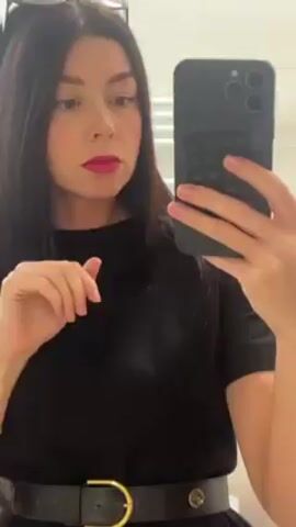 A woman is taking a selfie in a bathroom. She has long dark hair, pink lipstick, and a black outfit with a belt. Sunglasses are perched on her head. She is holding a black iPhone with a textured case. The bathroom has white tiles.