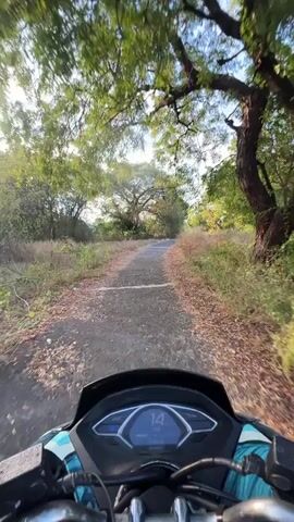 This is a first-person perspective video from a motorcycle traveling on a paved road. The road is surrounded by dry, yellowish vegetation and trees with green foliage. The sky is blue with some white clouds. The motorcycle's digital display shows speeds ranging from 15 to 26. The video captures a scenic drive through a rural or natural environment.