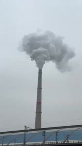 The video shows a tall industrial smokestack against a gray sky. A large plume of gray smoke rises from the top of the stack, spreading and swirling as it ascends. Below the smokestack, a blue and gray structure with what appears to be a series of vertical supports is visible. The overall impression is of an industrial setting with potential environmental impact.