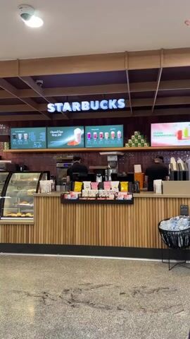 The video displays the interior of a Starbucks coffee shop, likely situated within a commercial complex like a mall or airport terminal in Nouaceur, Morocco. The space is brightly illuminated by artificial overhead lighting, and a security camera is mounted on the ceiling in the upper left.

Two male baristas, dressed in black shirts and caps, are actively working behind a long service counter. The counter features a distinctive front made of vertical wooden slats, topped with a light-colored surface. To the left, a glass display case showcases various pastries and baked goods, including croissants and muffins. The area behind the counter is equipped with coffee machines, blenders, and other preparation equipment.

Above the counter, a prominent "STARBUCKS" sign in white, illuminated letters is centered. Below it are three digital menu boards or promotional screens, primarily displaying text in Arabic. Visible Arabic phrases include "موا الهصيرات" (likely "Juice drinks" or "Drinks") and "اكثر روعة" ("more wonderful"). Another screen promotes "كراميل موكا" (Caramel Mocha) and "بالفراولة" ("with strawberry"). A larger digital display on the far right also shows promotional content.

In the foreground, to the right of the counter, there's a black, woven-design bin appearing to be for trash or recycling, containing several plastic water bottles. The floor is a mottled grey and brown, possibly tiled or concrete, with visible scuff marks. The overall atmosphere is clean and functional, with the baristas focused on their tasks.