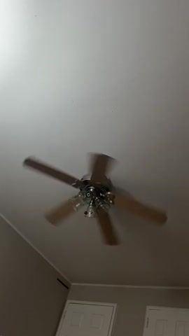 The image captures an upward perspective of an indoor room, dominated by a ceiling fan. The fan features five dark wood-toned or brown blades, which are in motion, indicated by motion blur. The central body of the fan and its mounting hardware are a metallic finish, possibly brushed nickel or chrome. Below the fan blades, an integrated light fixture is visible, consisting of three bell-shaped clear glass shades.

The ceiling is a plain white or off-white color. The upper portions of the walls visible in the frame are painted a neutral, light color, such as beige or light gray. A white trim or crown molding runs along the top edges of the walls where they meet the ceiling. On the left wall, a rectangular air vent or return air grille is present. In the lower part of the image, the tops of two white interior doors, each with two recessed panels, are partially visible, suggesting doorways or closet entrances.

A bright artificial light source, seemingly independent of the fan, illuminates the upper left corner of the ceiling. The overall lighting suggests an indoor setting, likely during the evening or night, or a room with limited natural light. No people or visible text are present in the image.