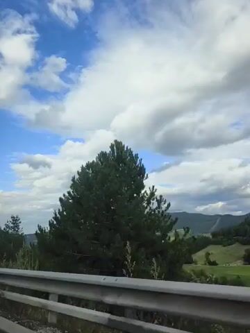 The video shows a view from a vehicle traveling along a road, with a guardrail in the foreground. The scene is a rural landscape with rolling hills covered in green vegetation and trees. The sky is blue with scattered white clouds. There are some trees on the right side of the road, some of which are very tall and appear to be pine trees. In the distance, the hills continue, and there are some scattered buildings or structures visible in the valleys. There is a traffic sign on the left side of the road, displaying a warning symbol with geometric shapes and a red triangle with a green light in the center. The overall impression is one of a scenic drive through a hilly, natural environment.