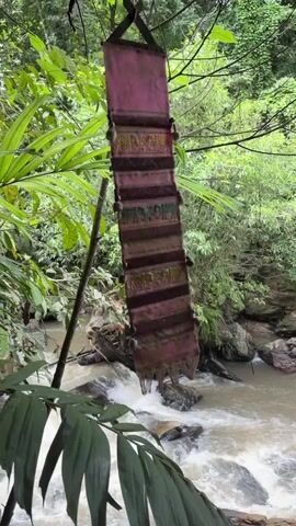 A hanging banner made of fabric is visible in a forest. The banner is composed of dark red and brown rectangular sections, with fringed edges at the bottom. It is suspended from a tree branch by two black straps forming an 'X' shape at the top. The banner sways gently in the breeze. Behind the banner, a stream flows over rocks, and lush green foliage surrounds the scene. The camera is positioned to capture the banner in the foreground with the stream and trees in the background.