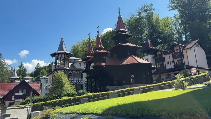 The video pans across a sunny street scene with traditional European-style buildings. In the foreground, a grassy slope leads up to several buildings with red-tiled roofs and dark wood accents. One prominent building resembles a church with multiple spires. The street is lined with cars and pedestrians, suggesting a moderately busy day. The architecture features steep roofs, balconies, and intricate detailing, characteristic of Alpine or Eastern European styles. Trees surround the buildings, indicating a natural, possibly mountainous, setting. The sky is clear blue with a few scattered clouds. The overall impression is of a charming, well-maintained town or resort area.