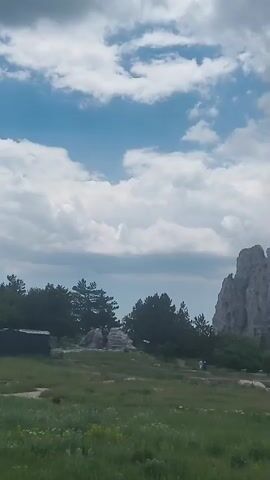 The video captures a scenic view of a rocky mountain range under a partly cloudy sky. In the foreground, a grassy meadow with scattered wildflowers stretches towards the mountains. To the left of the frame, a small, dark-colored structure, possibly a shed or cabin, is visible amongst some trees. A dirt path or road winds through the meadow and leads towards the mountainside. Two figures, appearing to be people walking, are seen on the path in the middle ground. The majestic mountain peaks dominate the right side of the frame, with a small red flag perched on the highest point of one of the prominent rock formations. Lush green trees and vegetation cover the slopes of the mountains, with a cable car line extending across the upper portion of the mountain range, suggesting a possible tourist attraction or transportation system. The sky is a blend of blue patches and large, fluffy white clouds. The overall impression is of a natural landscape, likely a park or a hiking destination, with elements of human presence indicated by the structures, path, and cable car.