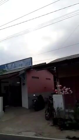This is a shaky, low-resolution video taken from a moving vehicle, likely a car, looking out the passenger side window. The video pans from right to left, showing a street scene. On the right side of the frame, a red building with a white trim is visible. A dark-colored motorcycle is parked in front of the building. Further left, a building with a white facade and a blue sign is seen, with a barber pole in front of it. A black scooter is parked to the left of the barber shop. Further down the street, more buildings and trees are visible, as well as power lines crisscrossing the sky. The sky is overcast. The overall impression is of a roadside view in a somewhat rural or suburban area, possibly in Southeast Asia, given the style of the buildings and vehicles.