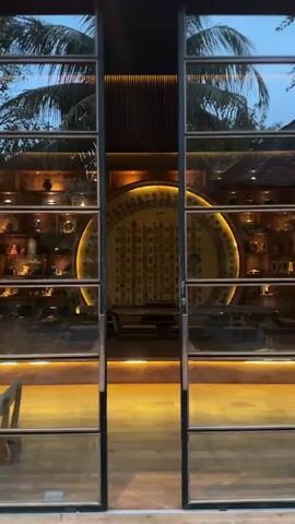 The video opens with a view through a large glass door, revealing an interior space with a distinctive circular wall feature. The circular feature is adorned with numerous Chinese characters and is illuminated by warm, indirect lighting, creating a dramatic focal point. The surrounding walls are lined with shelves displaying various decorative objects, likely ceramic vessels or similar items, also lit from above. The floor appears to be polished wood or a similar material, reflecting the ambient light. In the foreground, visible through the glass, are the frames of the door and a glimpse of outdoor foliage, suggesting palm trees, indicating a tropical or subtropical environment. As the camera moves forward and the doors open, the perspective shifts to directly face the interior. Several low, square seating cushions are arranged in rows in front of the circular wall art, suggesting a contemplative or dining area. The lighting throughout the space is dim and atmospheric, with a strong emphasis on warm tones, creating a sense of tranquility and sophistication. The overall impression is of a thoughtfully designed interior space with a strong cultural aesthetic.