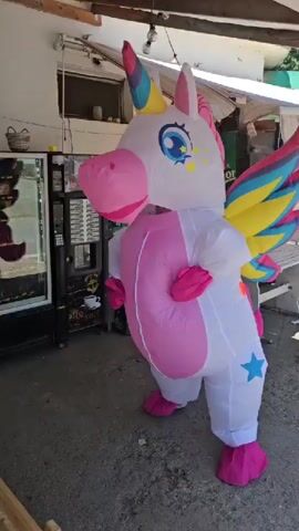 A person in a white inflatable unicorn costume with pink hooves, a pink tail, pink mane, and colorful wings stands facing left in an outdoor area with a concrete ground. Behind the unicorn is a vending machine and a white table with chairs. The unicorn costume has a blue star on its leg and an orange circle on its side. The person in the costume moves their arms in front of them.