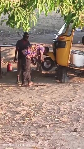 The video shows three men outdoors, standing around a yellow three-wheeled vehicle that is being worked on. One man, wearing a colorful shirt and dark pants, is bent over, appearing to work on the rear wheel. Another man, wearing a dark shirt and light-colored pants, stands watching. A third boy appears on a phone. The vehicle seems to be lifted by tires on the side. The ground is sandy and covered with debris, possibly leaves. A small red container sits nearby, and a table can be seen in the background. A tree with green leaves provides some shade. A pink motorbike passes the scene.